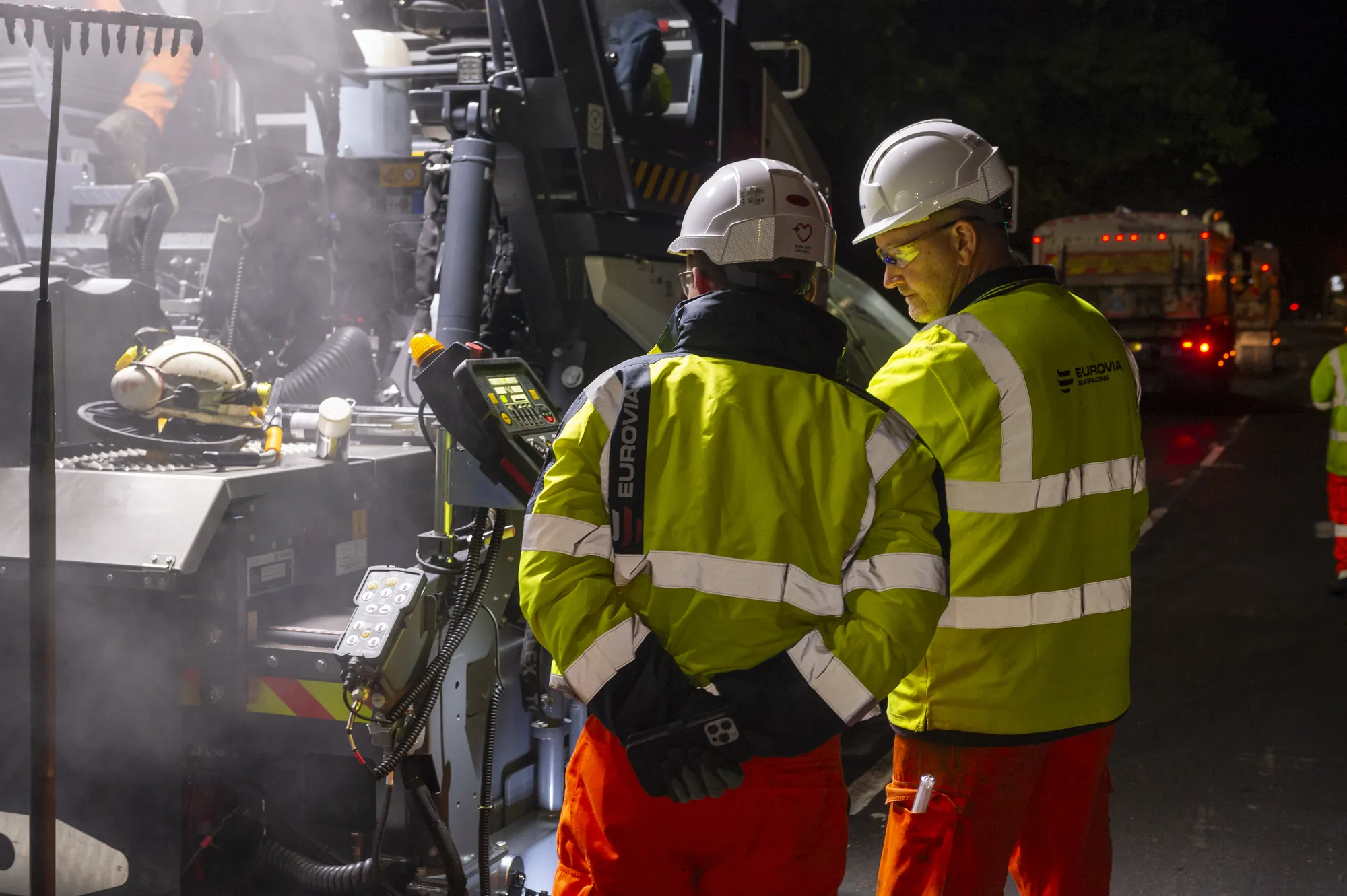Two Eurovia Surfacing operatives in helmets and high-vis clothing beside a paving machine at night