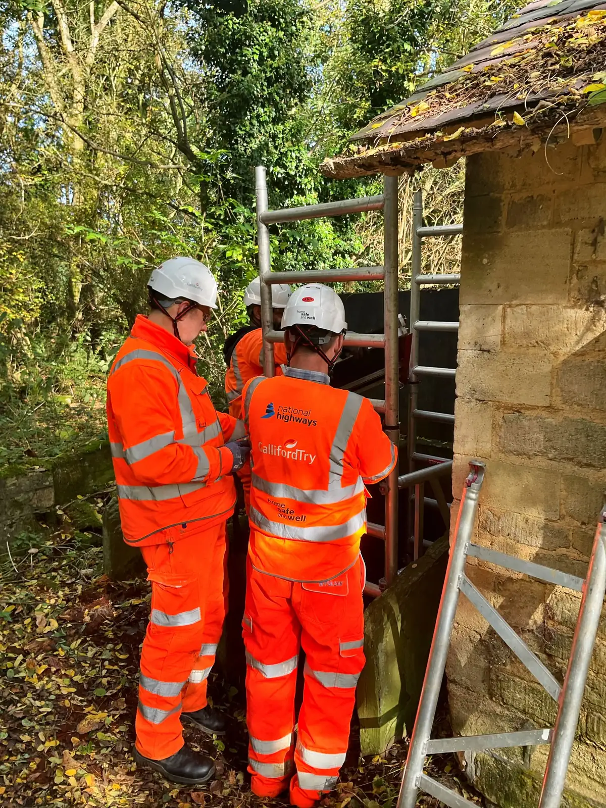 Two roadworkers in hi-vis inspecting a stone structure beside a ladder